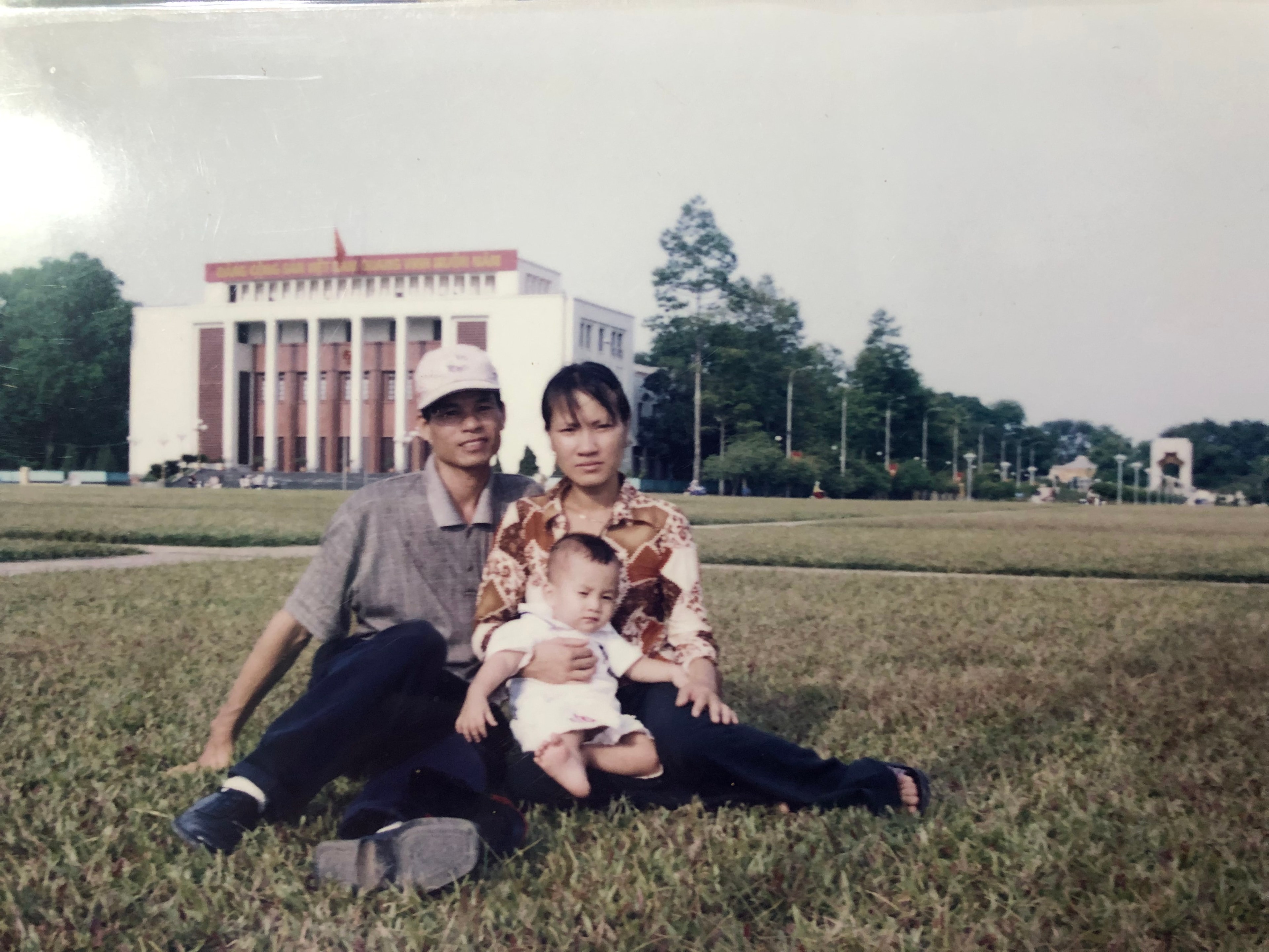 Two adults and a toddler sitting on the grass in front of Ho Chi Minh’s Mausoleum.
