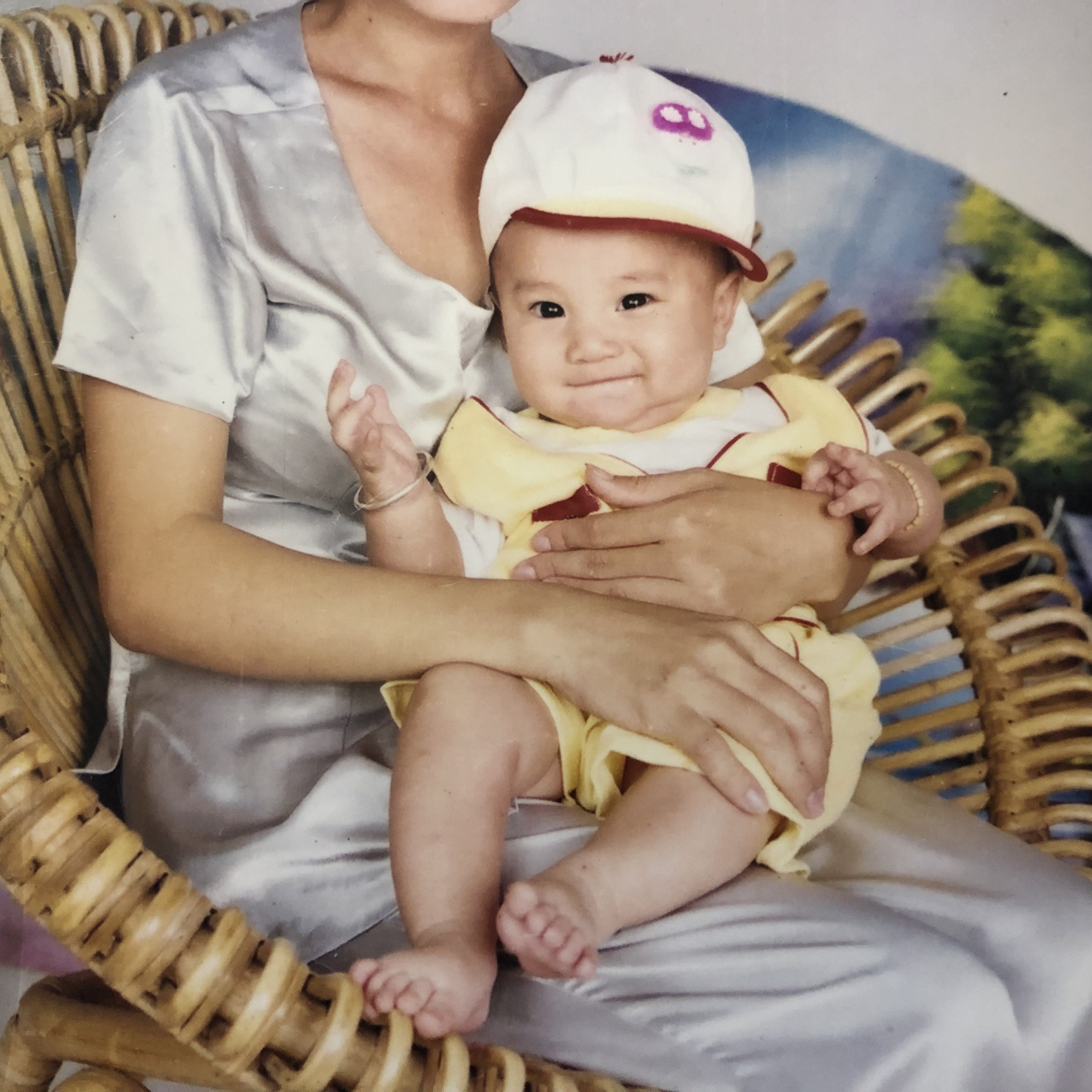 A baby in a yellow outfit and cap sitting on an adult’s lap in a wicker chair.