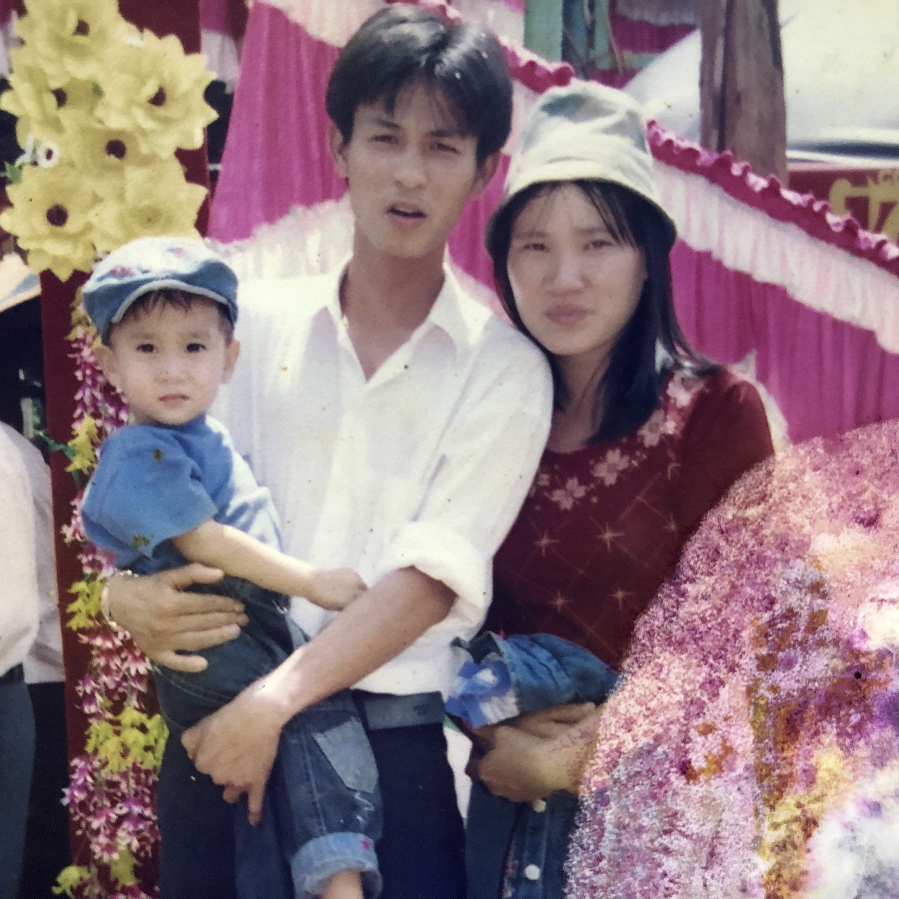A toddler held by an adult beside another adult, with wedding decorations behind them.